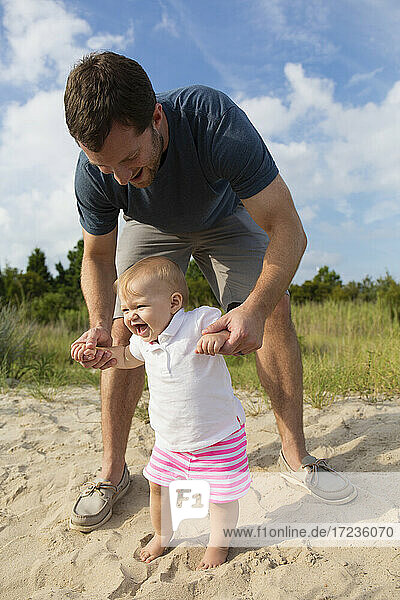 Mid adult man holding baby daughters hands while toddling in sand