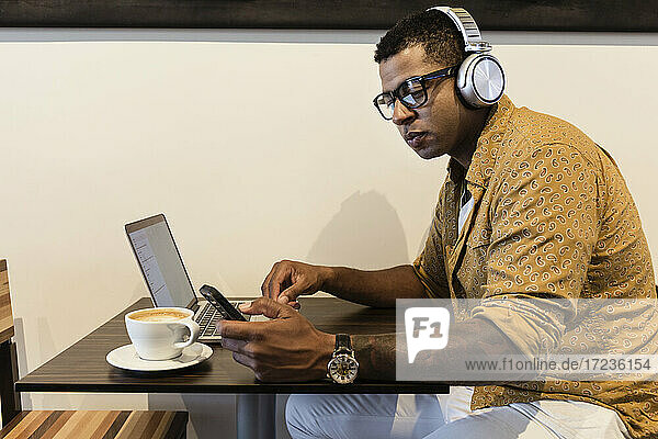 Young man in coffee shop  wearing headphones  using laptop and smartphone