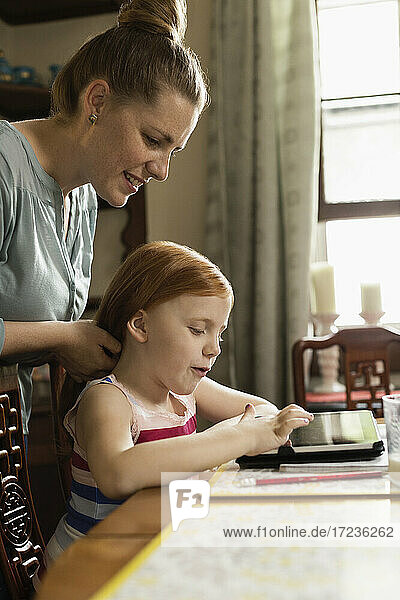 Mädchen mit digitalem Tablet am Esstisch  während die Mutter ihr Haar stylt