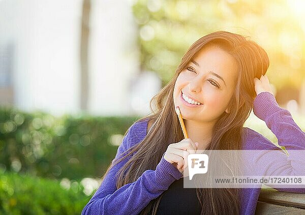 Attractive pensive mixed race female student with pencil sitting on campus bench