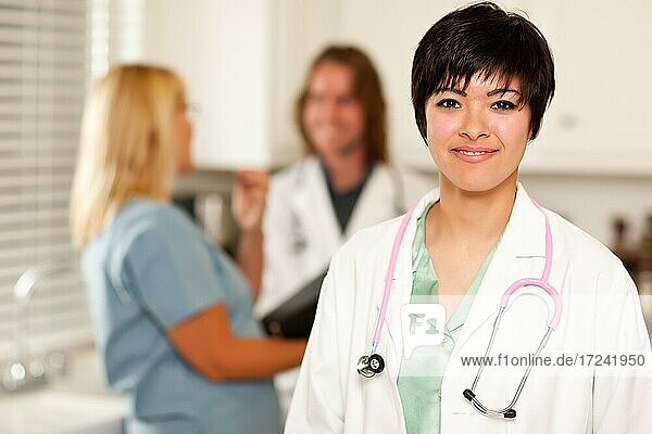 Hübsche Latino-Ärztin lächelt in die Kamera als Kollegen sprechen hinter ihr