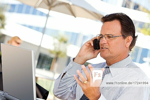 Gutaussehender Geschäftsmann  der mit seinem Handy telefoniert und im Freien am Laptop arbeitet