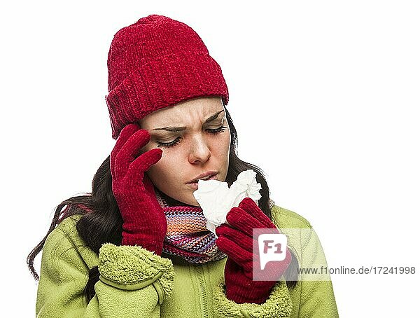 Kranke gemischtrassige Frau mit Wintermütze und Handschuhen schnäuzt sich die wunde Nase mit einem Taschentuch vor weißem Hintergrund