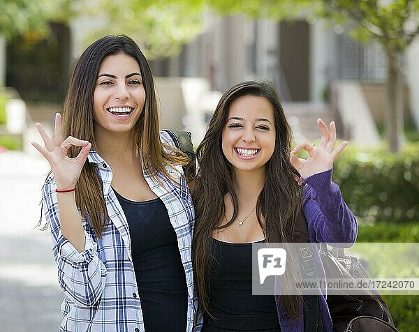 Portrait of two attractive mixed race female students with okay hand sign and carrying backpacks on school campus