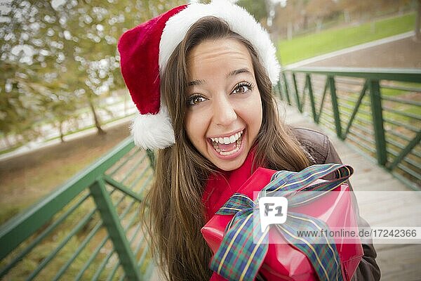 Hübsche festlich gekleidete lächelnde Frau trägt einen Weihnachtsmann Hut mit eingewickelten Geschenk außerhalb