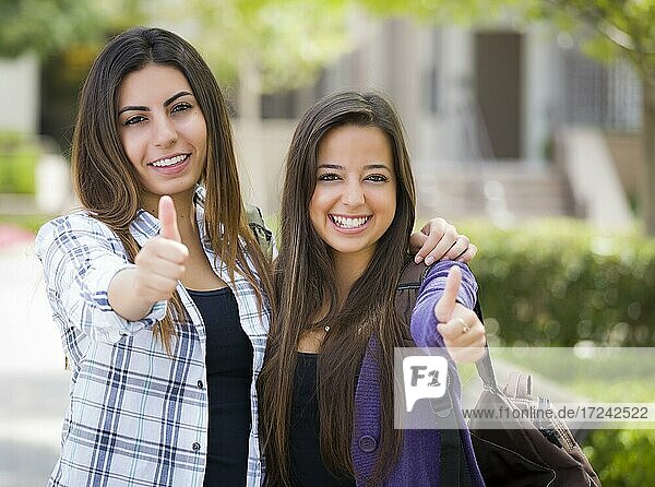 Porträt von zwei attraktiven multiethnischen Studentinnen mit Daumen nach oben und tragen Rucksäcke auf dem Schulcampus