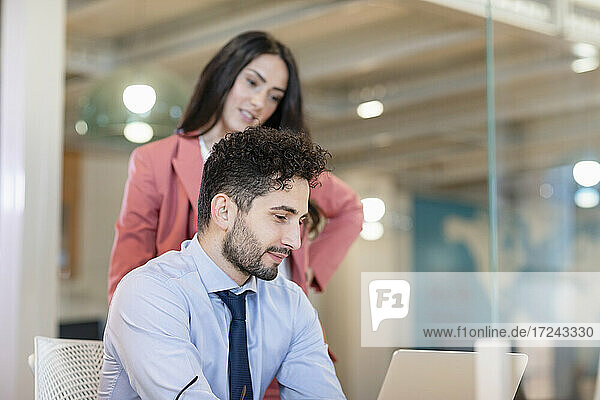 Geschäftsmann  der an einem Laptop arbeitet  während eine Kollegin in einem Coworking-Büro dahinter steht
