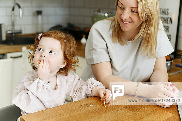 Smiling woman looking at daughter eating at dining table in kitchen