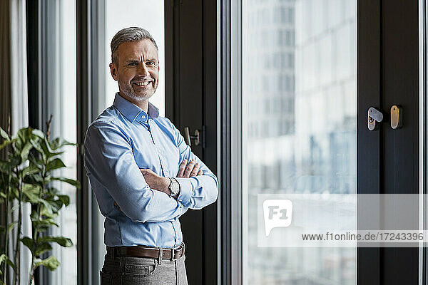 Smiling mature businessman with arms crossed standing by window at work place