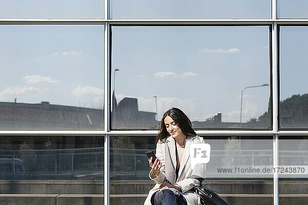 Geschäftsfrau  die ein Mobiltelefon benutzt  während sie vor einem Gebäude sitzt