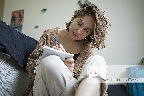 Frau mit braunen Haaren  die zu Hause vor dem Bett sitzt und auf ein Buch zeichnet