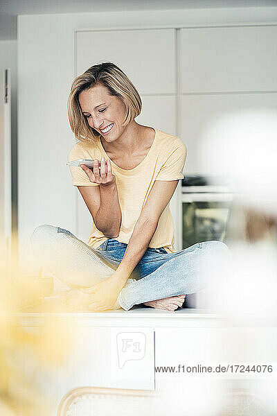 Lächelnde Frau mit gekreuzten Beinen  die zu Hause auf dem Küchentisch sitzt