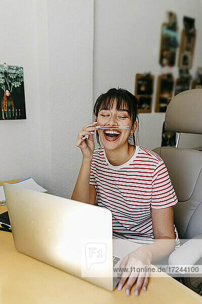 Fröhliche Frau  die mit einem Stift spielt  während sie mit einem Laptop im Büro zu Hause sitzt
