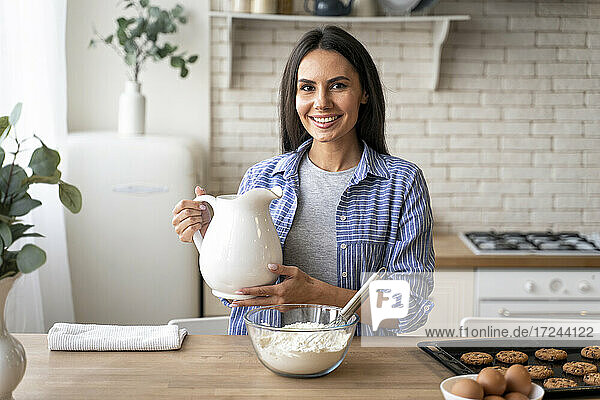 Lächelnde Frau hält Krug in der Nähe von Mehl in der Küche zu Hause