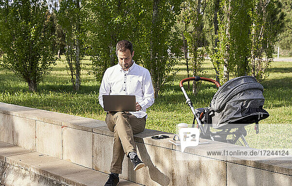 Mid adult businessman working on laptop while sitting by baby stroller in park