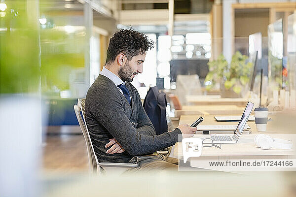 Porträt eines jungen Mannes mit Krawatte  der am Schreibtisch vor einem Laptop sitzt und auf sein Mobiltelefon schaut
