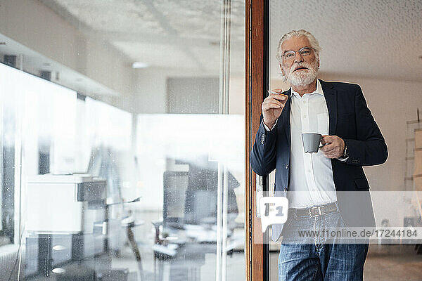 Älterer Geschäftsmann hält Zigarette mit Kaffeetasse  während er sich an eine Glaswand im Büro lehnt
