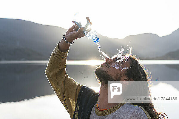 Mann gießt Wasser auf Gesicht mit Flasche in der Nähe von See während sonnigen Tag