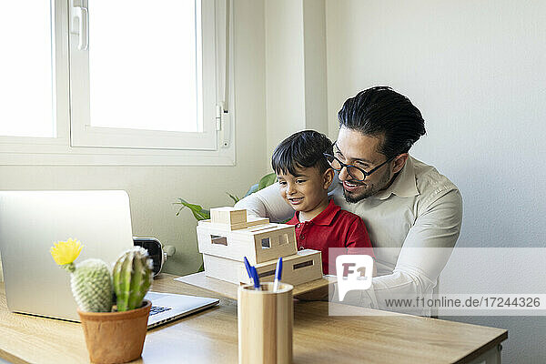 Männlicher Architekt  der ein Modell während eines Videogesprächs über einen Laptop zeigt  während er mit einem Jungen zu Hause im Büro sitzt
