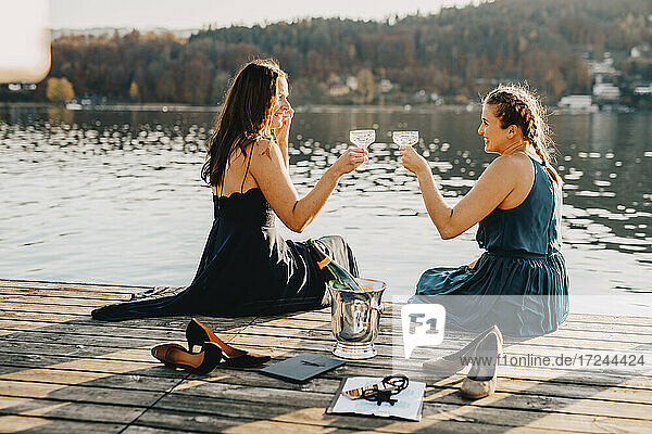 Weibliche Veranstaltungsplaner stoßen mit Champagner an  während sie auf dem Steg über dem See sitzen