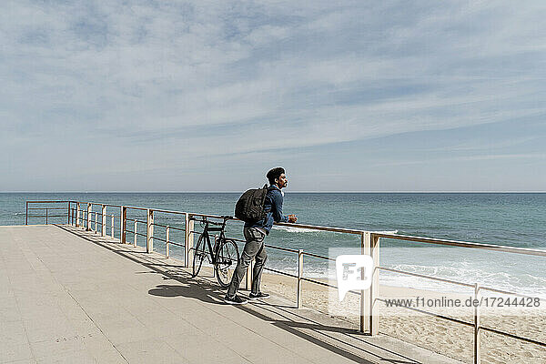 Mittlerer Erwachsener  der einen Rucksack trägt  während er sich mit dem Fahrrad am Strand auf das Geländer stützt  an einem sonnigen Tag
