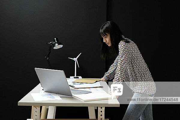 Geschäftsfrau  die auf einen Laptop schaut  während sie sich auf einen Schreibtisch an der Wand im Büro lehnt