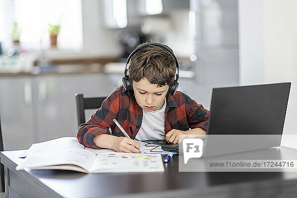 Boy wearing headphones writing on book while studying at home