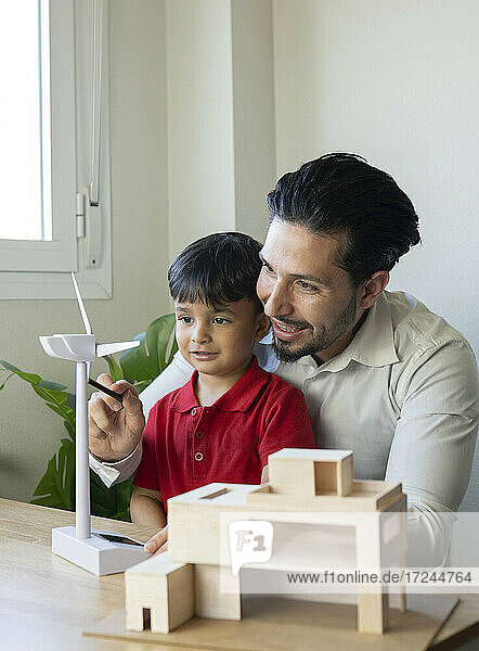 Männlicher Architekt  der das Modell einer Windkraftanlage untersucht  während er von seinem Sohn im Büro beobachtet wird