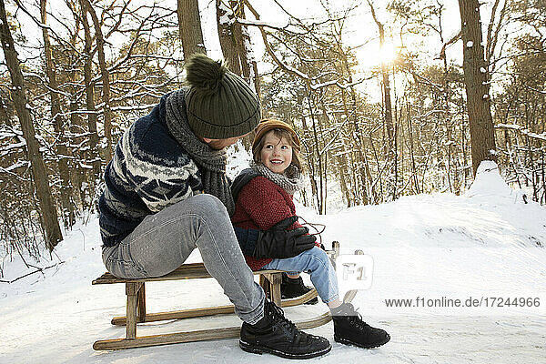 Father looking at son while sitting on sled during winter