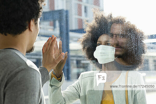Woman with protective face mask looking at male friend through glass wall