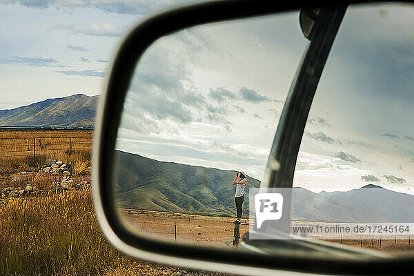 Side-view mirror reflection of young man standing on top of fence looking through binoculars at surrounding mountains