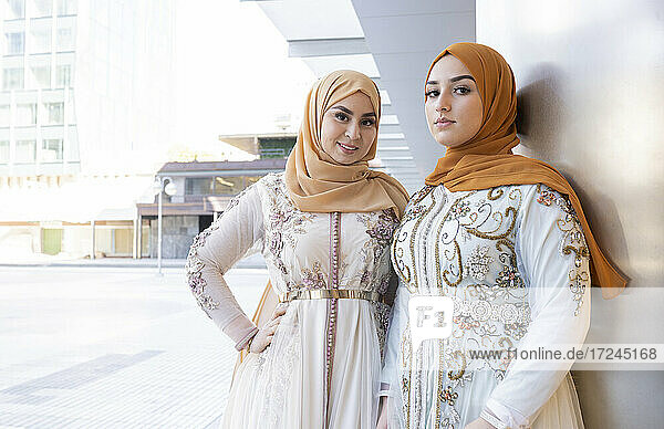 Beautiful friends in hijabs and dress standing together by wall
