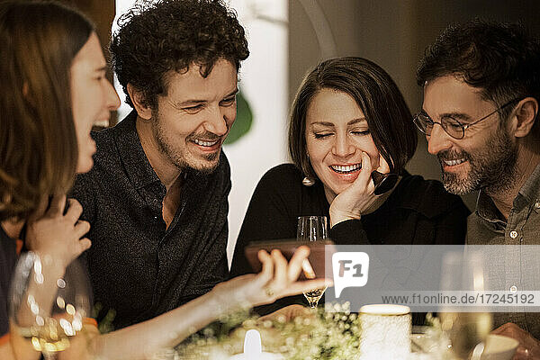 Fröhliche Frau zeigt Handy an lächelnde männliche und weibliche Freunde während einer Geburtstagsfeier zu Hause