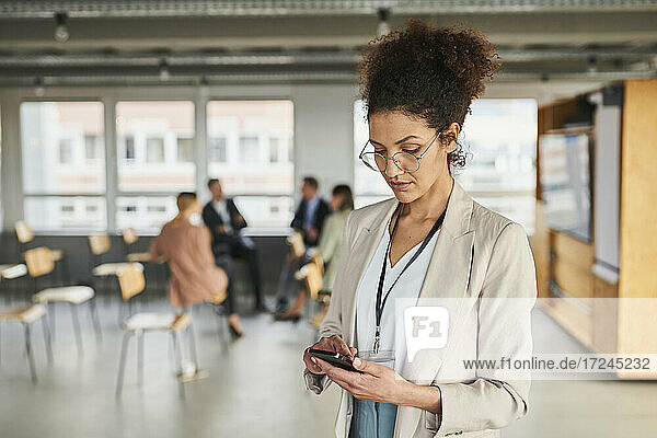 Unternehmerin bei der Nutzung eines Mobiltelefons mit Kollegen im Hintergrund im Büro