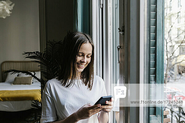 Lächelnde junge Frau benutzt ihr Smartphone am Fenster zu Hause