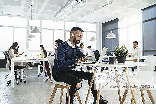 Männlicher Unternehmer arbeitet am Laptop mit Kollegen im Hintergrund in einem Coworking-Büro