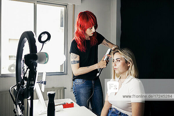 Weiblicher Friseur mit Glätteisen auf dem Haar des Modells vor Ringlicht im Studio
