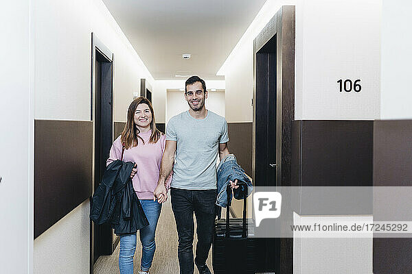 Lächelndes Paar  das sich an den Händen hält  während es mit Gepäck im Hotelflur spazieren geht