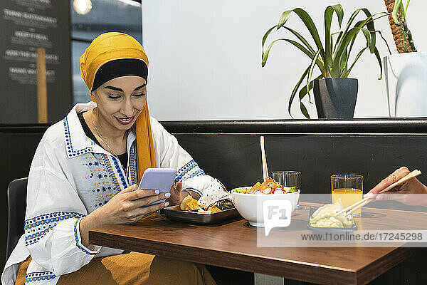 Young woman wearing headscarf using smart phone while sitting with female friend at restaurant