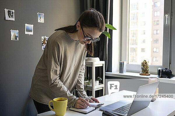 Junge Frau schreibt in ein Buch  während sie zu Hause am Laptop steht