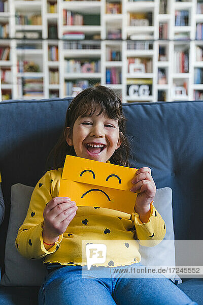 Lachendes Mädchen  das ein Emoticon mit traurigem Gesicht hält  während es auf dem Sofa zu Hause sitzt