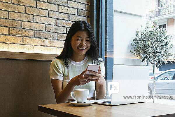 Lächelnde Frau  die ihr Smartphone am Laptop benutzt  während sie in einem Café sitzt