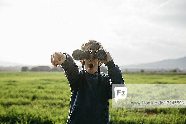 Surprised boy pointing while looking through binoculars in field during sunny day