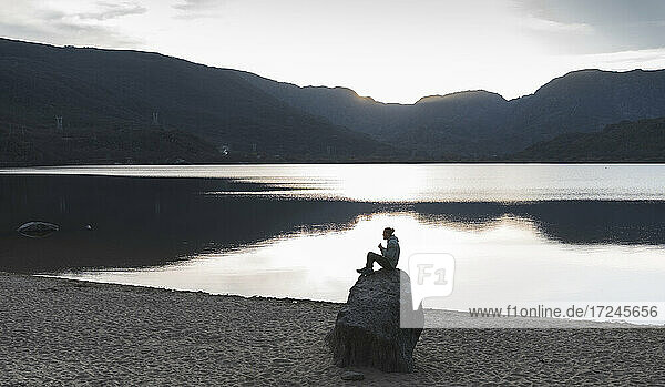 Mann sitzt bei Sonnenuntergang auf einem Felsen am See