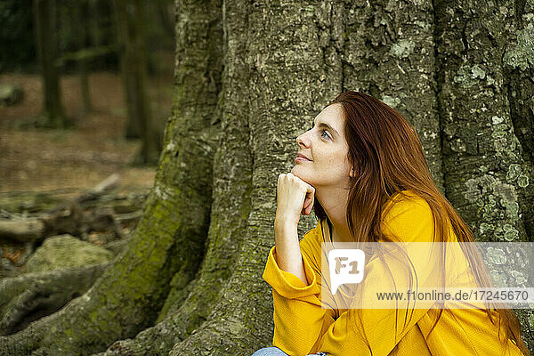 Young woman day dreaming by large tree trunk in forest