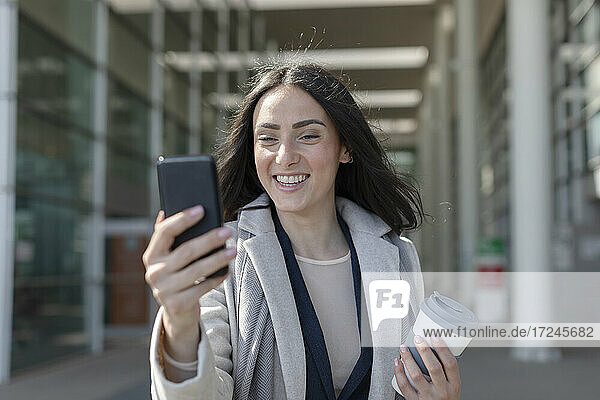 Lächelnde Geschäftsfrau mit Einwegbecher nimmt Selfie durch Smartphone