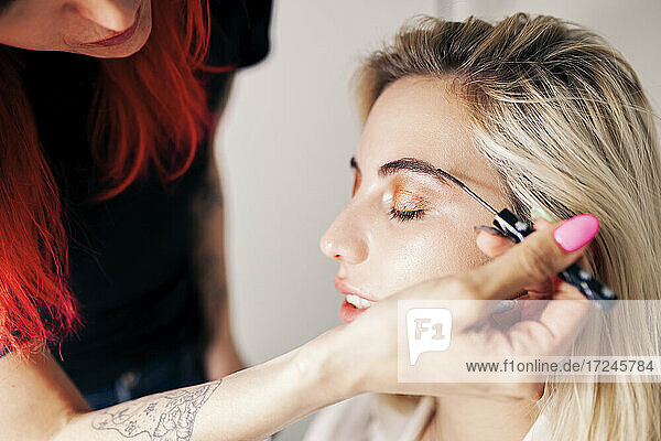 Young female make-up artist working on eyebrows of model with eyes closed at studio