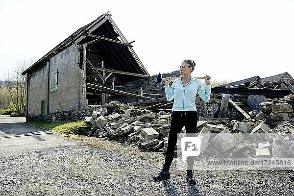 Thoughtful woman standing with hammer on road with destructed house in background