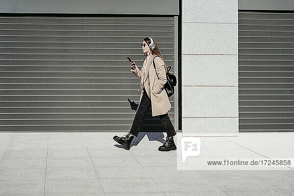 Junge Frau mit Kopfhörern und Mobiltelefon auf dem Gehweg