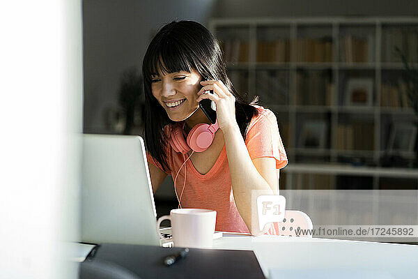 Lächelnde Geschäftsfrau  die mit einem Smartphone spricht  während sie mit einem Laptop im Büro zu Hause sitzt